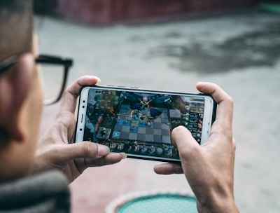 A person is holding a smartphone horizontally, immersed in playing a complex strategy game. The focus is on the game screen, which displays an intricate board setup with various characters and elements.